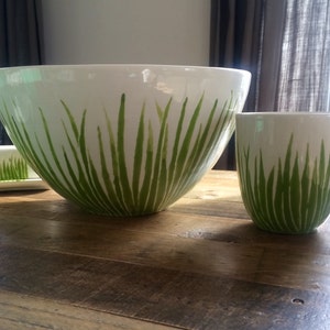 May include: A white ceramic bowl and mug with a green grass design. The bowl is large and the mug is smaller. Both are sitting on a wooden table.