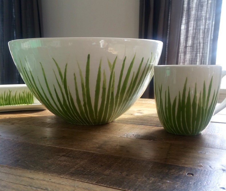 May include: A white ceramic bowl and mug with a green grass design. The bowl is large and the mug is smaller. Both are sitting on a wooden table.