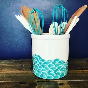May include: White ceramic utensil holder with a teal and blue scale pattern. The holder is filled with wooden spoons and teal whisks.
