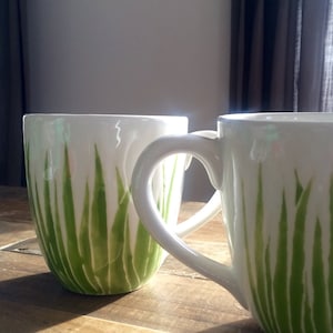 May include: Two white ceramic mugs with green grass designs. The mugs have heart-shaped handles.