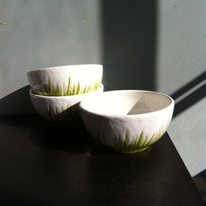 May include: Three white ceramic bowls with a green grass design. The bowls are stacked on top of each other on a black surface.