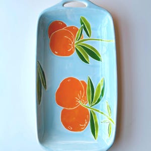 May include: A light blue ceramic serving dish with two orange fruit designs. The dish has two handles on the sides.