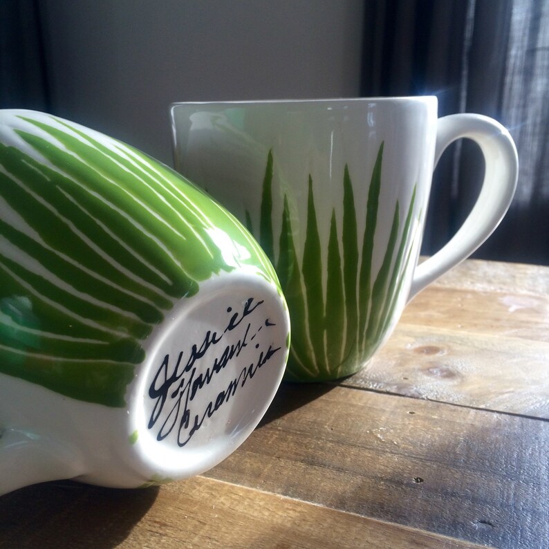 May include: Two white ceramic mugs with green leaf designs. The mug on the left has the words "Jessie Javanil Ceramics" written on the bottom in black.
