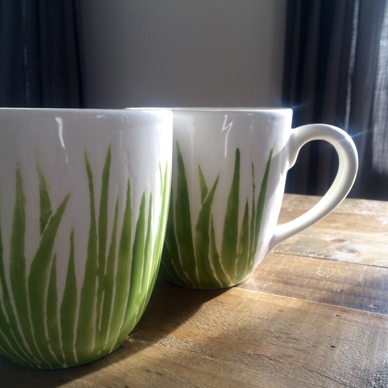 May include: Two white ceramic mugs with green grass designs. The mugs are on a wooden surface.