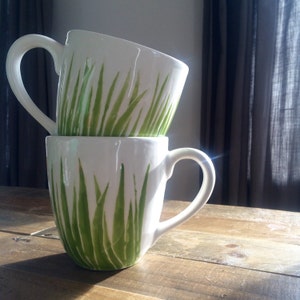 May include: Two white ceramic mugs with green grass designs stacked on top of each other. The mugs have a handle on the right side.