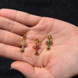 May include: Three gold-coloured cross pendants, each adorned with colourful gemstones. One cross features clear stones, another has pink and purple stones, and the third has green stones. The pendants are displayed on a hand.
