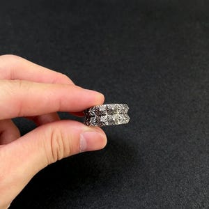 Puede incluir: Un anillo plateado con un patrón floral repetido. El anillo se sostiene entre dos dedos sobre un fondo oscuro. El diseño del anillo presenta una serie de pequeñas formas simétricas en forma de flor, creando una superficie texturizada.
