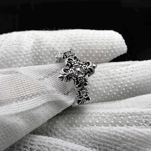 Puede incluir: Un anillo plateado con un diseño de cruz detallado. El anillo presenta patrones ornamentados y un elemento de flor de lis en la parte superior de la cruz. El anillo se muestra sobre una superficie blanca texturizada.