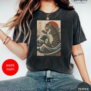 May include: Dark gray t-shirt featuring a Japanese-inspired graphic of a cat monster in a wave. The graphic includes a red sun and Japanese text. The shirt is a crew neck and has short sleeves.