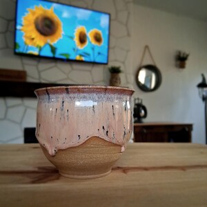 May include: A handmade ceramic bowl with a light pink glaze and brown rim. The glaze has dark drips and a textured, unglazed base. The bowl sits on a wooden surface, with a television displaying sunflowers in the background.