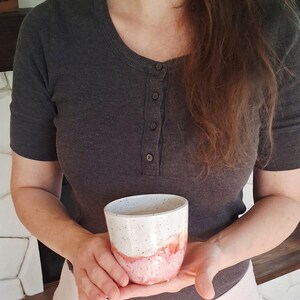 May include: A handmade ceramic mug with a white speckled top and a pink and orange watercolor design on the bottom half. The mug is held in front of a person wearing a gray shirt and a light pink skirt.