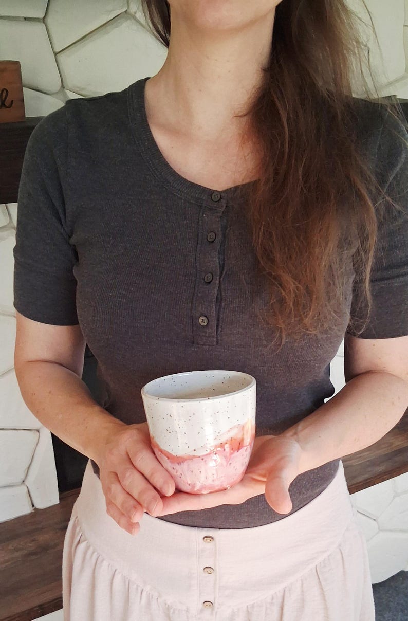 May include: A handmade ceramic mug with a white speckled top and a pink and orange watercolor design on the bottom half. The mug is held in front of a person wearing a gray shirt and a light pink skirt.