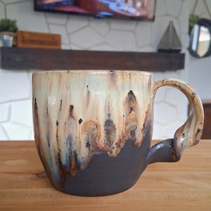 May include: A handmade ceramic mug with a unique design. The mug features a dark gray base that transitions to a cream and brown upper section with blue accents. The handle has a distinctive, sculpted shape. This mug is perfect for coffee or tea.