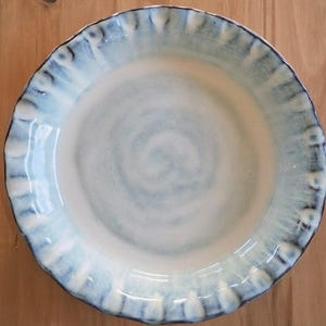 May include: A round ceramic bowl with a smooth, off-white center and a wide, scalloped rim featuring a gradient of light blue and dark blue brushstrokes. The interior surface displays a subtle, swirling texture, and the edges are defined by dark blue.
