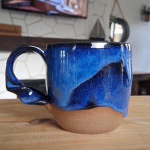 May include: A handmade ceramic mug with a twisted handle. The mug features a two-tone design, with a deep blue glaze on the upper portion and a natural brown base. The mug is sitting on a wooden surface.