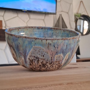 May include: A round ceramic bowl featuring a reactive glaze with swirling shades of blue, light purple, and cream. The exterior and interior surfaces display a mottled texture with dark brown speckles and drips cascading down the sides.