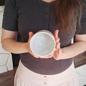 May include: A speckled ceramic bowl with a light gray exterior and interior. The bowl is held in both hands, showcasing its round shape and speckled design. The bowl appears to be handmade.