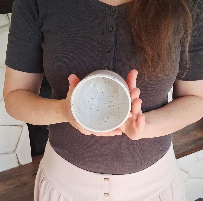 May include: A speckled ceramic bowl with a light gray exterior and interior. The bowl is held in both hands, showcasing its round shape and speckled design. The bowl appears to be handmade.