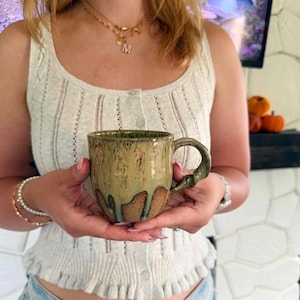May include: A handmade ceramic mug with a unique glaze. The mug features a green and brown color scheme with a dripped glaze effect. The handle is also green. The mug is held in front of a white top.