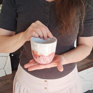 May include: A speckled ceramic cup with a white top, transitioning to a peach and pink band, and a speckled pink base. The cup is held in a person's hands, showcasing its handmade quality and unique design.