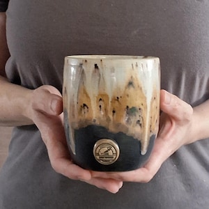 May include: A ceramic mug with a cylindrical shape, featuring a two-tone glaze. The upper portion is a creamy off-white with amber-colored drips, while the lower section is a solid matte black. A circular tan medallion with text is centered.