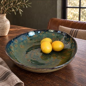 May include: A large, shallow, round ceramic bowl with a glossy, mottled glaze in shades of deep blue, forest green, and brown. Three bright yellow lemons are resting inside the bowl, which features an irregular, organic rim and a smooth finish.