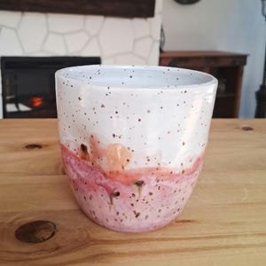 May include: A handmade ceramic mug with a speckled white upper half and a pink and peach-colored lower half. The mug has a rounded shape and is likely used for drinking beverages. The mug is sitting on a wooden surface.