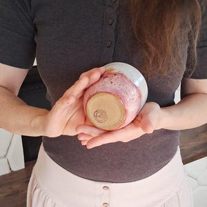 May include: A handmade ceramic bowl with a speckled pink and white glaze. The bowl is held in two hands, showcasing its rounded shape and unique color variations. The bottom of the bowl has a stamped design.