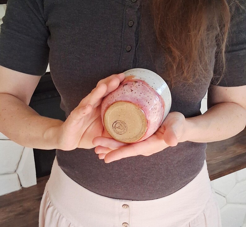 May include: A handmade ceramic bowl with a speckled pink and white glaze. The bowl is held in two hands, showcasing its rounded shape and unique color variations. The bottom of the bowl has a stamped design.
