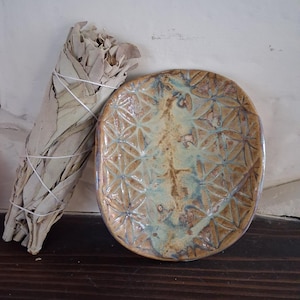 May include: An oval ceramic dish with a geometric pattern in shades of brown, tan, and blue. A bundle of dried sage is next to the dish. The sage is wrapped with white string. The items are on a dark wooden surface.