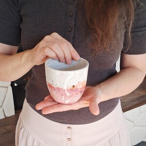 May include: A handmade ceramic mug with a white speckled top and a pink and peach watercolor design on the bottom half. The mug is held in a person's hands, showcasing its unique, artistic design. This mug is perfect for coffee, tea, or any beverage.