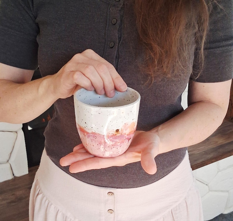 May include: A handmade ceramic mug with a white speckled top and a pink and peach watercolor design on the bottom half. The mug is held in a person's hands, showcasing its unique, artistic design. This mug is perfect for coffee, tea, or any beverage.