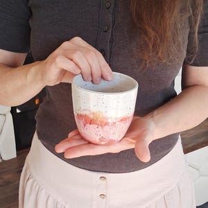 May include: A handmade ceramic cup with a speckled white top, transitioning to a pink and orange watercolor design at the base. The cup is held in both hands, showcasing its unique, artistic design. The cup is perfect for coffee or tea.