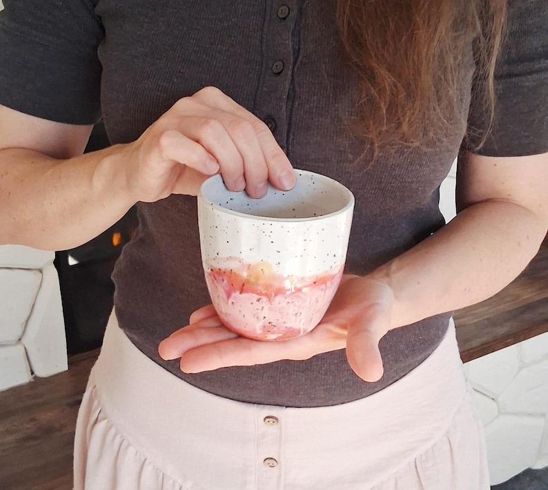 May include: A handmade ceramic cup with a speckled white top, transitioning to a pink and orange watercolor design at the base. The cup is held in both hands, showcasing its unique, artistic design. The cup is perfect for coffee or tea.