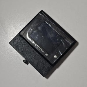 Puede incluir: Un dispositivo LCD Smart Battery negro con una pantalla cubierta de plástico. El dispositivo está en una funda negra texturizada. El texto "SMART BATTERY LCD" está impreso en el dispositivo.