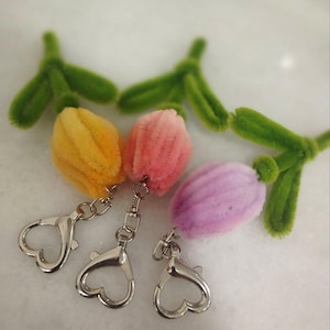 May include: Three handmade tulip keychains in yellow, pink, and purple. Each tulip has a green stem and leaves made of fuzzy pipe cleaners. The keychains have silver-coloured heart-shaped clasps.