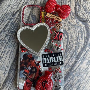 May include: A clear phone case decorated with a large heart-shaped mirror, a red rhinestone bow, a money bag charm, and a Parental Advisory Explicit Content label. It features a photo of a person in a red dress, a cross charm, and red accents.