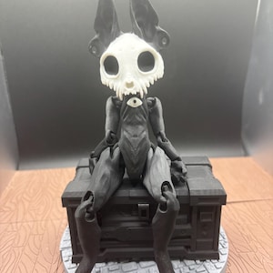 May include: A black and white articulated figurine of a creature with a skull-like head and large ears. The figurine is seated on a dark gray chest, with a round base. The head is white, and the body is dark gray.