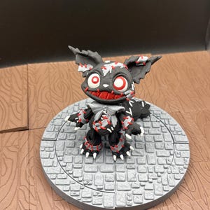 Puede incluir: Una figura de criatura negra y gris con detalles rojos, incluyendo ojos y costuras. La figura tiene orejas grandes y está posada sobre una plataforma circular gris con un diseño de adoquines. El tema general es un diseño espeluznante inspirado en Halloween.