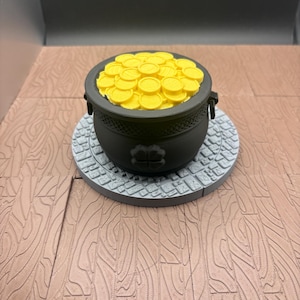 May include: A black pot filled with yellow coins, resting on a gray stone-like base. The pot has two handles and a four-leaf clover design. The base sits on a wooden surface.