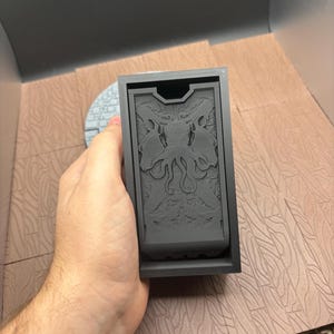 Puede incluir: Una caja rectangular gris oscuro con un detallado relieve de una criatura con tentáculos y alas. La caja se sostiene en una mano, mostrando el intrincado diseño. La caja parece ser un contenedor de almacenamiento.