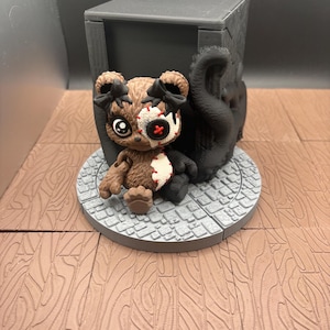 May include: A brown and black teddy bear figurine with a stitched eye and a red cross. The bear sits on a gray circular platform with a dark gray backdrop. The platform is on a brown wooden surface.