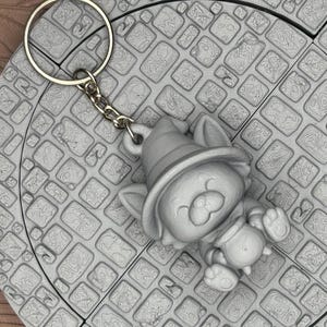 May include: A gray keychain featuring a cartoon cat wearing a witch hat and holding a cauldron. The cat is smiling. The keychain is attached to a silver chain and ring. The background is a gray, textured surface.