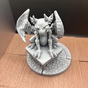 May include: A gray gargoyle figurine with detailed wings, horns, and clawed feet. The statue is perched on a square base atop a circular platform with a cobblestone design. The figurine is a fantasy-themed decorative item.