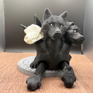 Puede incluir: Figura negra de perro de tres cabezas con un cráneo unido a una cabeza. La figura tiene detalles de pelaje y patas, y está sobre una base circular. Los ojos de las cabezas tienen una cualidad reflectante.