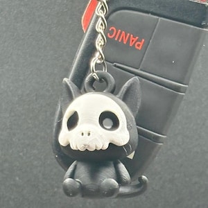 May include: A black keychain featuring a small, stylized cat-like figure with a skull-like face. The keychain is attached to a car key fob with a red accent and the word "PANIC" visible.