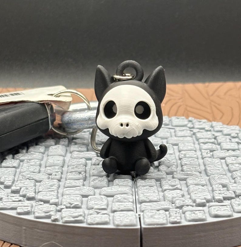 May include: A black and white keychain featuring a cartoon cat with a skull-like face. The cat has large, round eyes and is sitting on a gray, textured surface. A set of keys is visible in the background.