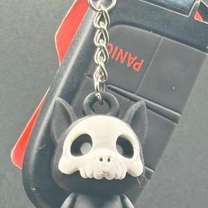 May include: A black and white keychain featuring a small, stylized dog with a skull-like face. The keychain is attached to a car key fob with the word "PANIC" visible.