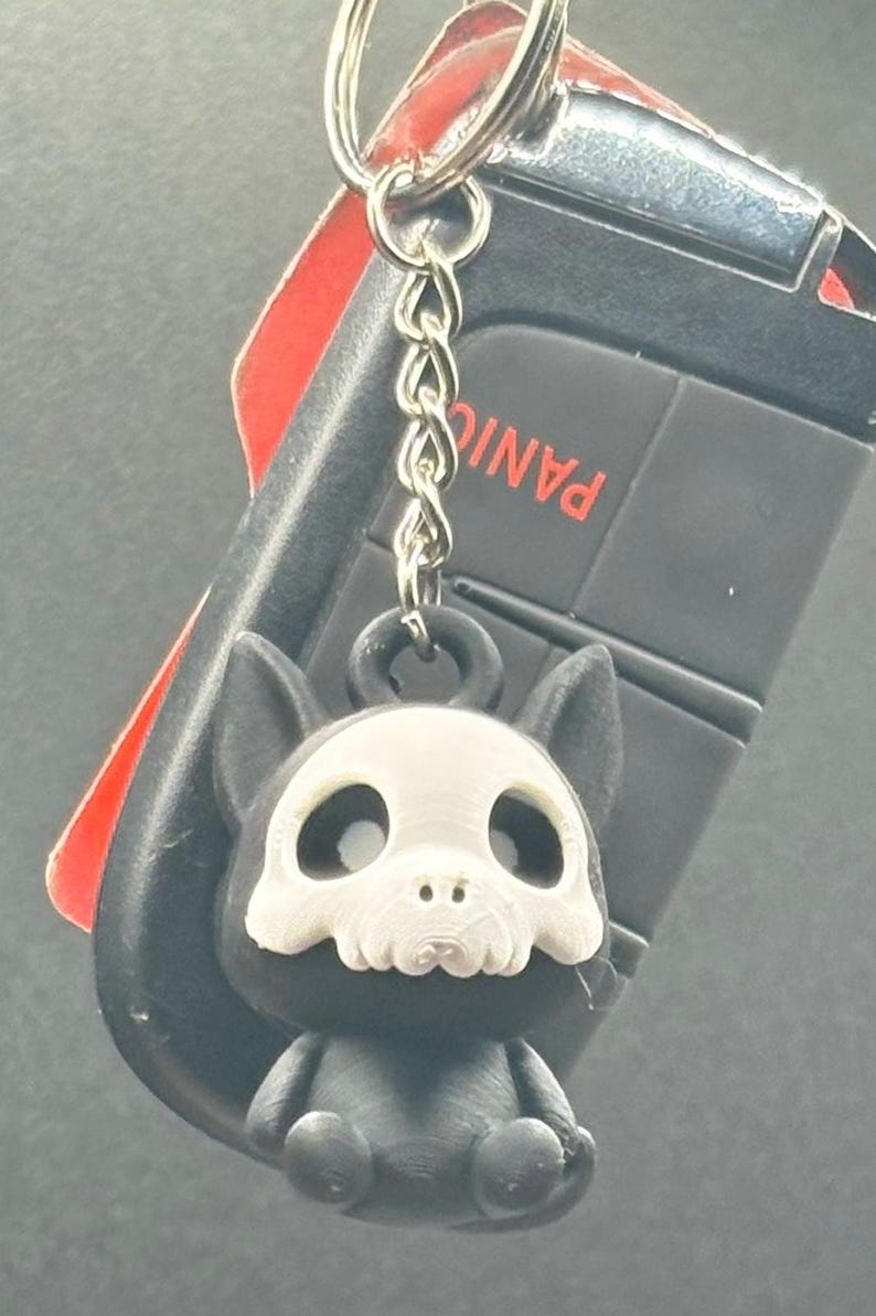 May include: A black and white keychain featuring a small, stylized dog with a skull-like face. The keychain is attached to a car key fob with the word "PANIC" visible.