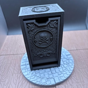 May include: A matte black rectangular storage box featuring a skull and crossbones emblem on the front panel. The box has intricate wood-grain textures and decorative scrollwork, and it sits on a circular base with a cobblestone pattern.
